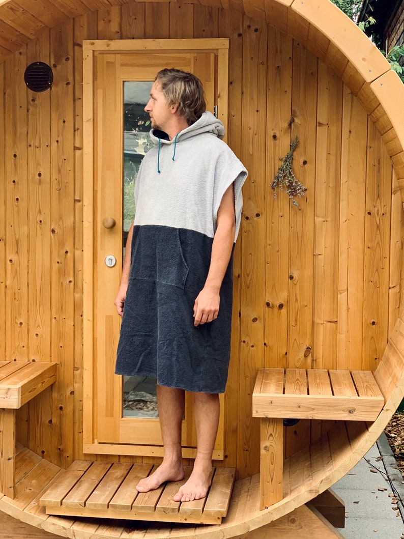 May include: A person wearing a grey and black hooded towel robe stands in front of a wooden sauna. The sauna has a wooden door and two benches.