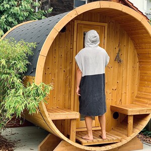 May include: A wooden barrel sauna with a person standing in the doorway wearing a grey and black hooded towel robe. The sauna has a wooden bench and a small window.
