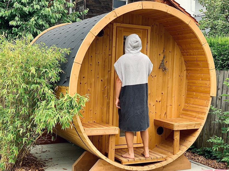 May include: A wooden barrel sauna with a person standing in the doorway wearing a grey and black hooded towel robe. The sauna has a wooden bench and a small window.