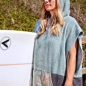 K&ouml;nnte beinhalten: Ein hellblaues Kapuzenhandtuch mit einer Tasche mit Kartenmotiv. Das Handtuch wird von einer Person getragen, die ein Surfbrett h&auml;lt.