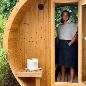 May include: A wooden barrel sauna with a person standing in the doorway wearing a grey hooded towel. The sauna has a wooden bench and a white bucket with a wooden ladle.