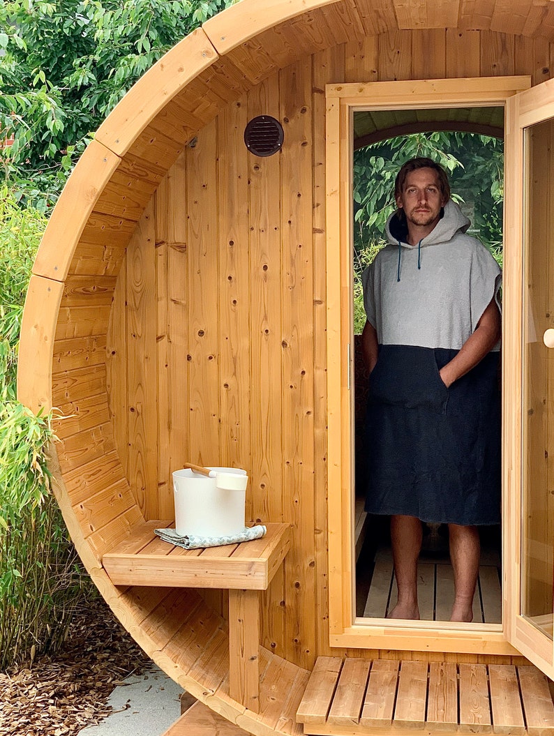 May include: A wooden barrel sauna with a person standing in the doorway wearing a grey hooded towel. The sauna has a wooden bench and a white bucket with a wooden ladle.