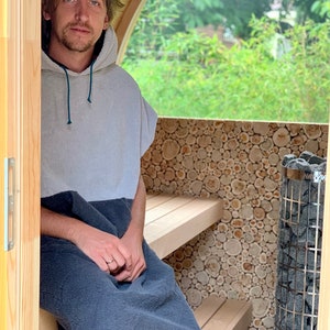 May include: A person wearing a grey and blue hooded towel sits on a wooden bench inside a small wooden sauna. The sauna has a wood-burning stove with a black metal door and a window that looks out onto a green garden.