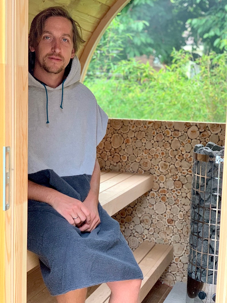 May include: A person wearing a grey and blue hooded towel sits on a wooden bench inside a small wooden sauna. The sauna has a wood-burning stove with a black metal door and a window that looks out onto a green garden.
