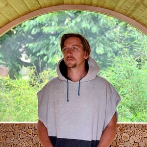May include: A person wearing a grey hooded towel with a blue drawstring. The person is standing in a wooden structure with a large arched window behind them.