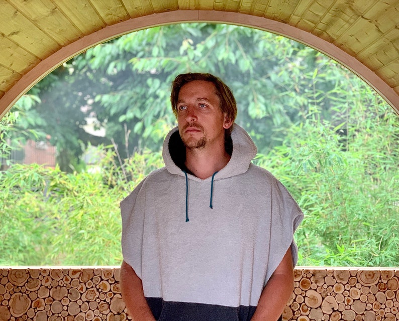 May include: A person wearing a grey hooded towel with a blue drawstring. The person is standing in a wooden structure with a large arched window behind them.