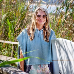 K&ouml;nnte beinhalten: Eine Frau mit Sonnenbrille und einem hellblauen Kapuzenhandtuch h&auml;lt ein wei&szlig;es Surfbrett vor einem Hintergrund aus gr&uuml;nen Schilf.