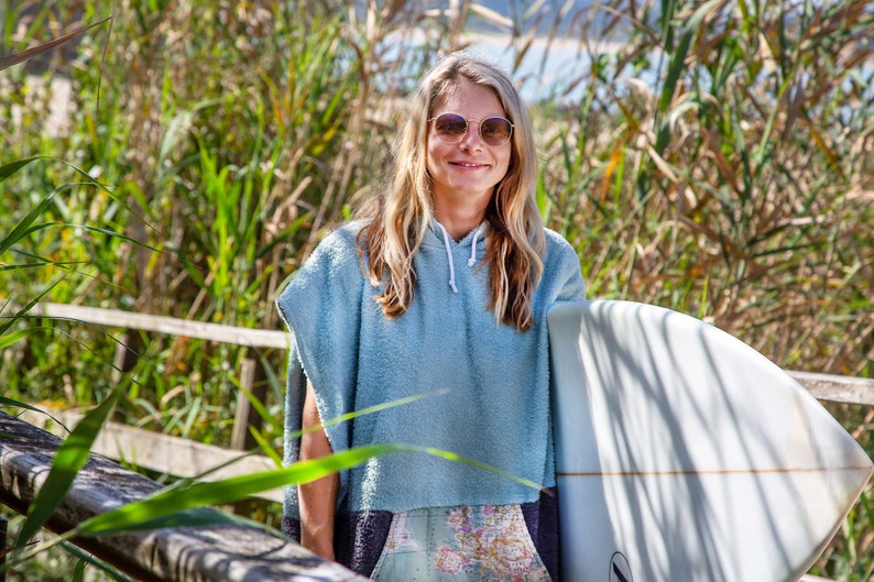 K&ouml;nnte beinhalten: Eine Frau mit Sonnenbrille und einem hellblauen Kapuzenhandtuch h&auml;lt ein wei&szlig;es Surfbrett vor einem Hintergrund aus gr&uuml;nen Schilf.