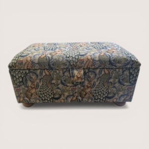 May include: A rectangular ottoman with a fabric cover featuring a repeating pattern of rabbits, peacocks, and foliage in shades of blue, green, and brown. The ottoman rests on four small, rounded wooden feet.