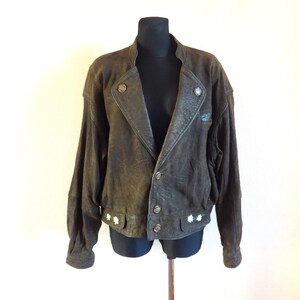 Puede incluir: Una chaqueta de cuero marr&oacute;n con cierre de cremallera y detalles de botones. La chaqueta tiene cuello y mangas largas. Hay flores blancas bordadas en la parte inferior de la chaqueta.