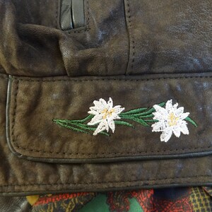 Puede incluir: Primer plano de una chaqueta de cuero marr&oacute;n con un bolsillo que presenta flores de edelweiss blancas bordadas.