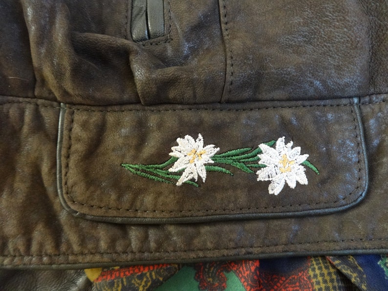 Puede incluir: Primer plano de una chaqueta de cuero marr&oacute;n con un bolsillo que presenta flores de edelweiss blancas bordadas.