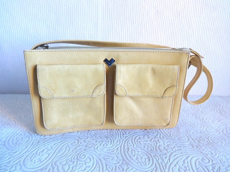 May include: A light yellow leather shoulder bag with two large front pockets and a small metal heart-shaped logo.
