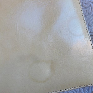 May include: A light beige leather bag with a stitched edge and a strap. The bag has two light-colored stains on the front.