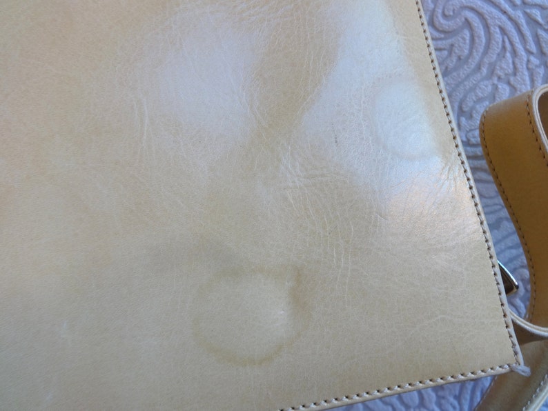May include: A light beige leather bag with a stitched edge and a strap. The bag has two light-colored stains on the front.