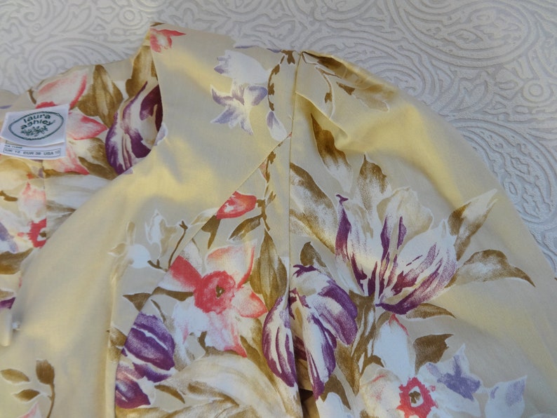 Puede incluir: Primer plano de una prenda color crema con estampado floral. El dise&ntilde;o presenta flores grandes en tonos morado, rosa y marr&oacute;n. La prenda tiene una etiqueta que dice "laura ashley".