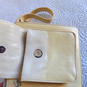 May include: A light beige leather crossbody bag with a flap closure and two silver metal snaps. The bag has a small pocket on the front.