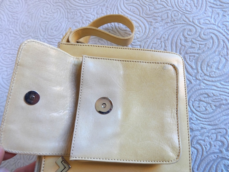 May include: A light beige leather crossbody bag with a flap closure and two silver metal snaps. The bag has a small pocket on the front.