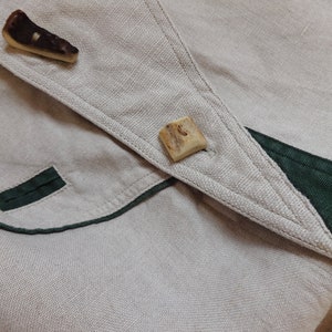 Pu&ograve; includere: Primo piano di un capo in lino beige con finiture verde scuro e due esclusivi bottoni in corno. Il tessuto ha un aspetto strutturato e i bottoni sono di forma irregolare.