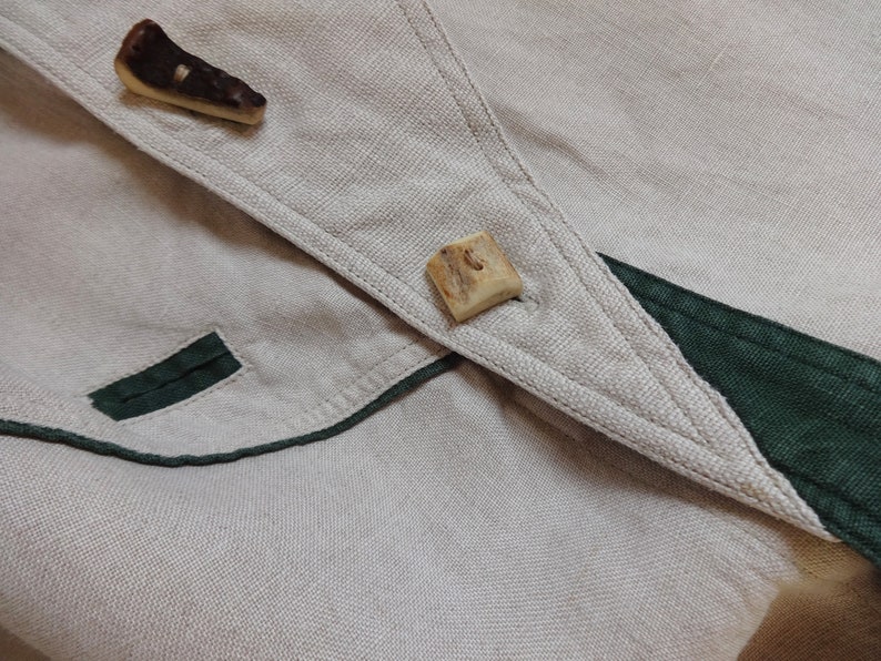 Pu&ograve; includere: Primo piano di un capo in lino beige con finiture verde scuro e due esclusivi bottoni in corno. Il tessuto ha un aspetto strutturato e i bottoni sono di forma irregolare.