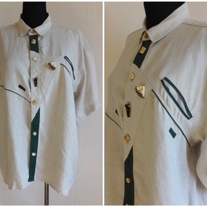 Pu&ograve; includere: Camicia beige a maniche corte con dettagli verde scuro e neri. La camicia presenta un'abbottonatura frontale con bottoni decorativi e disegni geometrici. Il colletto e il pannello frontale hanno finiture verde scuro.