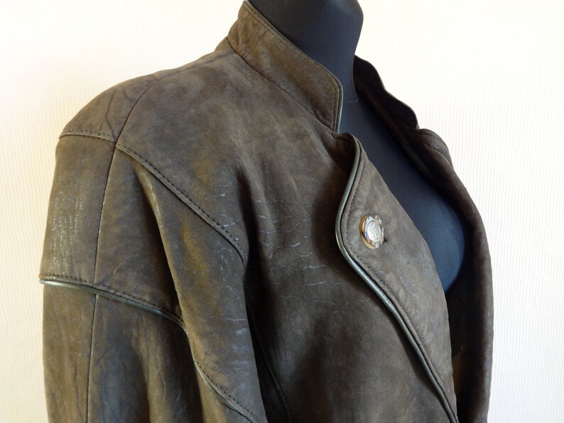 Puede incluir: Una chaqueta de cuero marr&oacute;n con cuello mandar&iacute;n y un cierre de bot&oacute;n &uacute;nico. La chaqueta tiene un aspecto ligeramente desgastado y presenta un bot&oacute;n decorativo en la parte delantera.