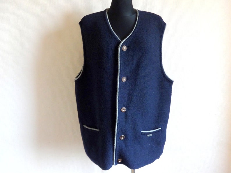Puede incluir: Un chaleco de punto azul marino con ribete gris alrededor del cuello, las sisas y los bolsillos. El chaleco tiene cierre de botones y dos bolsillos.