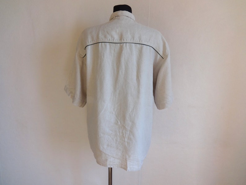 Pu&ograve; includere: Camicia di lino beige chiaro con maniche corte, vista da dietro. La camicia ha un colletto e un bordo nero sulle spalle e sulla parte superiore della schiena. Il tessuto sembra leggero e traspirante.