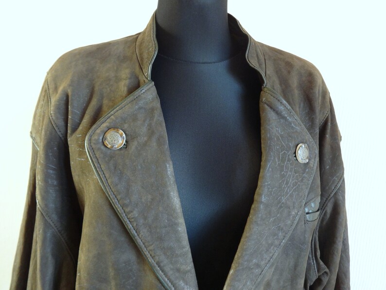 Puede incluir: Una chaqueta de cuero marr&oacute;n con cuello alto y dos botones plateados. La chaqueta tiene un aspecto desgastado y est&aacute; hecha de cuero suave y flexible.