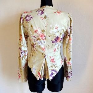 Puede incluir: Una blazer beige con estampado floral y mangas largas, exhibida en un maniqu&iacute;. La chaqueta presenta un patr&oacute;n de flores rosas y moradas con hojas verdes. La parte trasera de la blazer tiene un corte entallado con una abertura en la cintura.