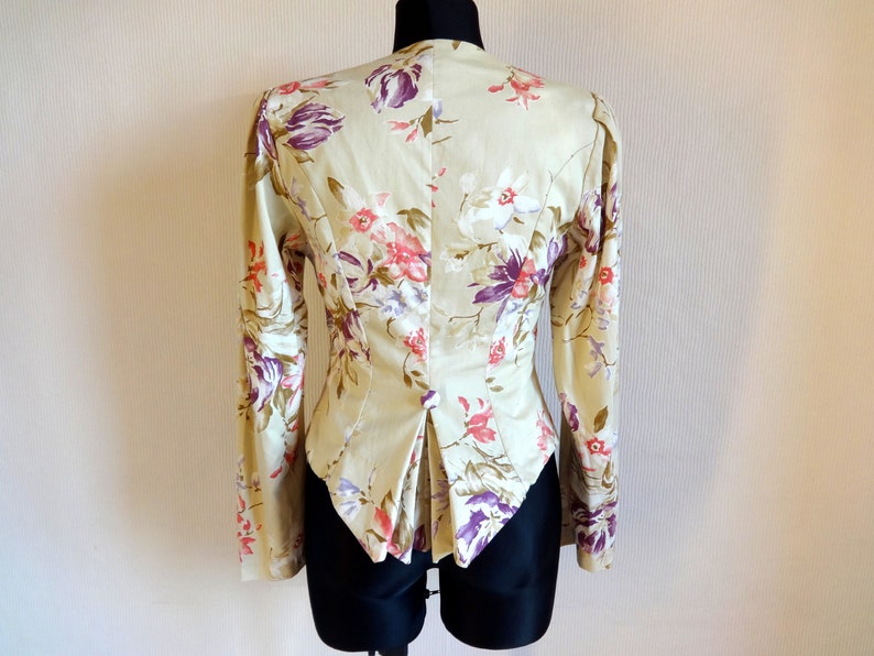 Puede incluir: Una blazer beige con estampado floral y mangas largas, exhibida en un maniqu&iacute;. La chaqueta presenta un patr&oacute;n de flores rosas y moradas con hojas verdes. La parte trasera de la blazer tiene un corte entallado con una abertura en la cintura.