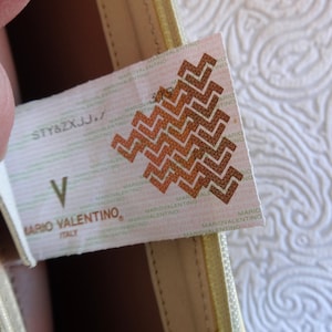 May include: A white tag with the text "STY&ZXJJ." and "MARIO VALENTINO, ITALY" printed on it. The tag also features a brown geometric pattern.