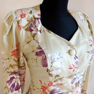 Puede incluir: Blazer beige floral con cuello con muescas y tres botones grandes y redondos. La tela presenta un estampado de flores moradas, rosas y blancas con hojas verdes. El blazer tiene mangas largas y hombros acolchados.