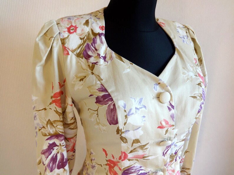 Puede incluir: Blazer beige floral con cuello con muescas y tres botones grandes y redondos. La tela presenta un estampado de flores moradas, rosas y blancas con hojas verdes. El blazer tiene mangas largas y hombros acolchados.