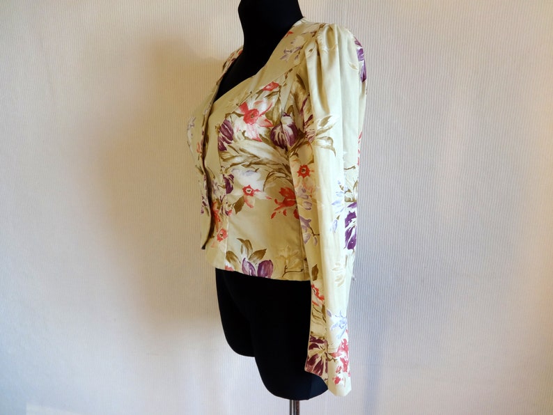 Puede incluir: Chaqueta verde claro con estampado floral y mangas largas. La chaqueta presenta un escote en V y est&aacute; adornada con un patr&oacute;n de flores rojas, moradas y blancas. La prenda se exhibe en un maniqu&iacute; negro.
