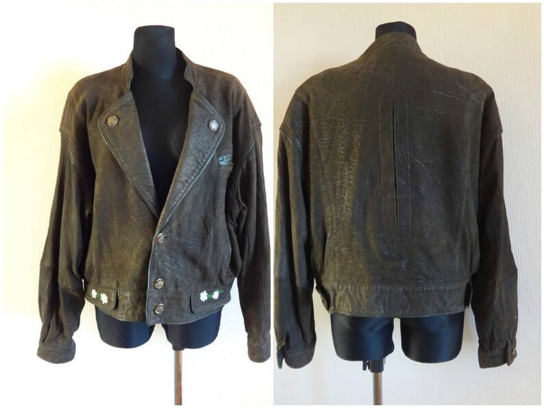 Puede incluir: Una chaqueta de cuero marr&oacute;n con cierre de cremallera y detalles de botones. La chaqueta tiene un aspecto desgastado y presenta flores blancas bordadas en el dobladillo inferior.