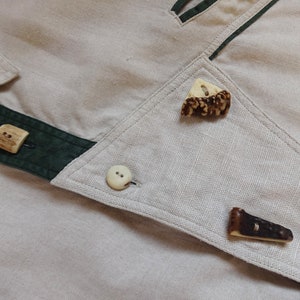 Pu&ograve; includere: Primo piano di un capo in lino beige con finiture verde scuro e bottoni decorativi. I bottoni sono realizzati con materiali naturali, tra cui corna e ossa, aggiungendo un tocco rustico e unico al design.