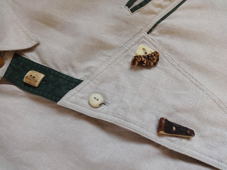 Pu&ograve; includere: Primo piano di un capo in lino beige con finiture verde scuro e bottoni decorativi. I bottoni sono realizzati con materiali naturali, tra cui corna e ossa, aggiungendo un tocco rustico e unico al design.