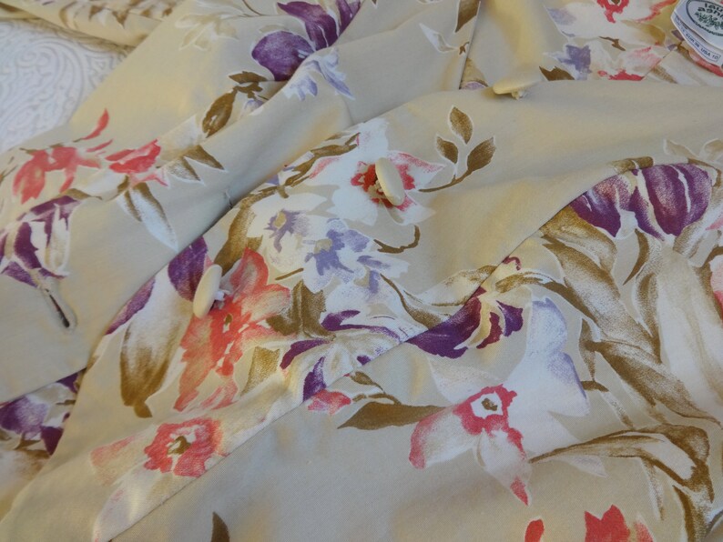 Puede incluir: Tela beige con un estampado floral con flores rosas, moradas y blancas y hojas marrones. La tela est&aacute; drapeada, mostrando la textura y el dise&ntilde;o. La prenda tiene botones blancos.