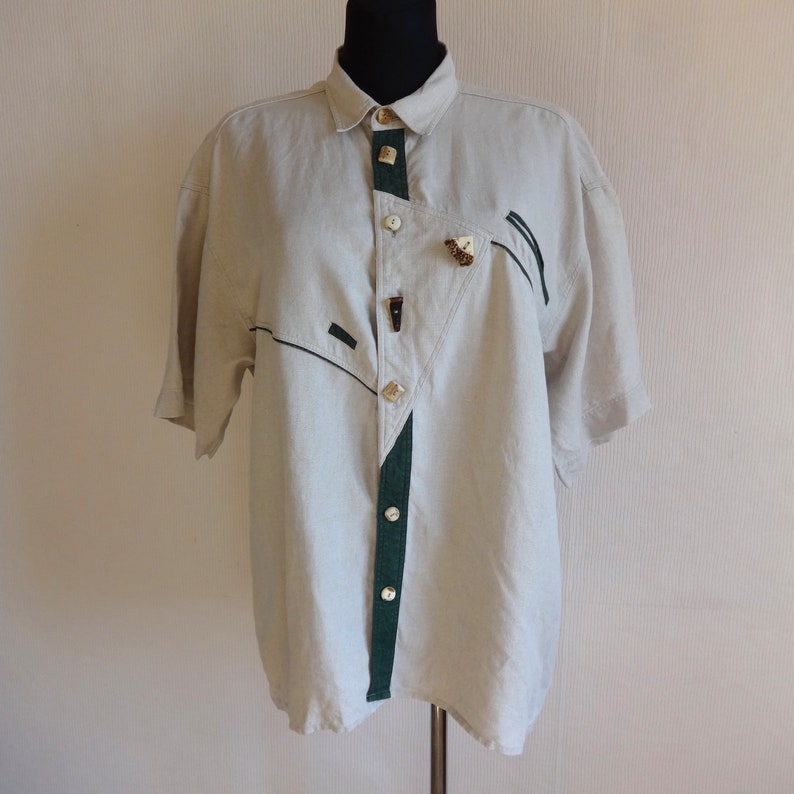 Pu&ograve; includere: Camicia beige chiaro a maniche corte con un design unico. La camicia presenta dettagli verde scuro, bottoni decorativi e un motivo geometrico. Il colletto e i bottoni sono di colori contrastanti. La camicia &egrave; esposta su un manichino.