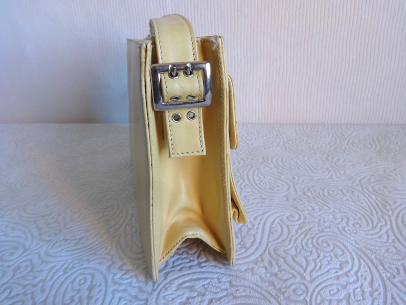 May include: A pale yellow leather shoulder bag with a silver buckle and strap. The bag has a flap closure and a structured shape.