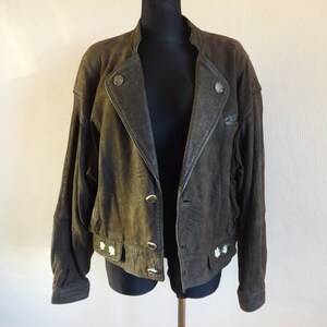 Puede incluir: Una chaqueta de cuero verde oscuro con cierre de cremallera y detalles de botones. La chaqueta tiene cuello y mangas largas. Hay flores blancas bordadas en la parte inferior de la chaqueta.