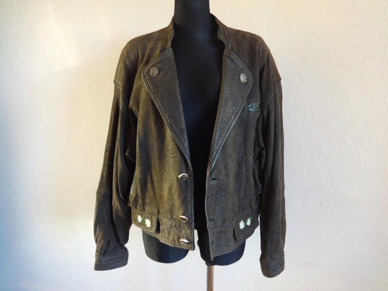 Puede incluir: Una chaqueta de cuero verde oscuro con cierre de cremallera y detalles de botones. La chaqueta tiene cuello y mangas largas. Hay flores blancas bordadas en la parte inferior de la chaqueta.