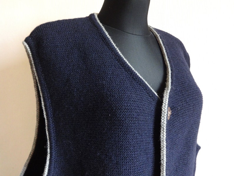 Puede incluir: Un chaleco de punto azul marino con ribete gris alrededor del cuello y la abertura de botones. El chaleco tiene cuello en V y un solo cierre de bot&oacute;n.