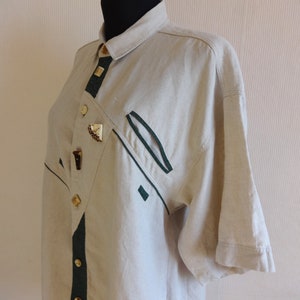 Pu&ograve; includere: Camicia di lino beige a maniche corte. La camicia presenta finiture verde scuro e bottoni decorativi. Il design include un motivo asimmetrico unico con una tasca ed elementi decorativi. La camicia &egrave; esposta su un manichino nero.