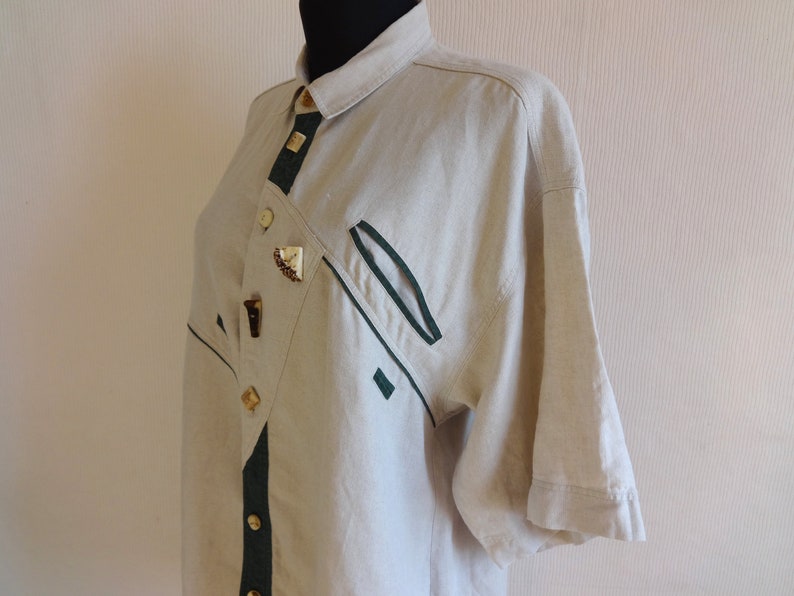 Pu&ograve; includere: Camicia di lino beige a maniche corte. La camicia presenta finiture verde scuro e bottoni decorativi. Il design include un motivo asimmetrico unico con una tasca ed elementi decorativi. La camicia &egrave; esposta su un manichino nero.