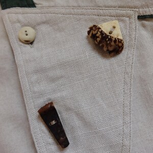 Pu&ograve; includere: Primo piano di un tessuto beige chiaro con tre bottoni. Uno &egrave; un bottone rotondo di colore chiaro. Gli altri due sono bottoni unici, marroni e beige, dalla forma irregolare. Il tessuto ha un bordo verde.