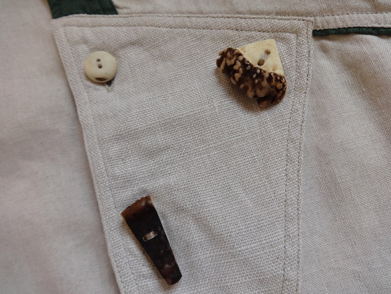 Pu&ograve; includere: Primo piano di un tessuto beige chiaro con tre bottoni. Uno &egrave; un bottone rotondo di colore chiaro. Gli altri due sono bottoni unici, marroni e beige, dalla forma irregolare. Il tessuto ha un bordo verde.