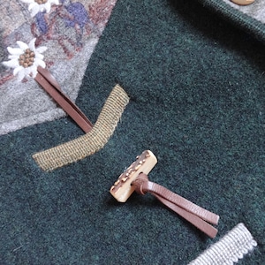 Puede incluir: Primer plano de una chaqueta de lana verde con cierre de bot&oacute;n de cuero marr&oacute;n. El bot&oacute;n est&aacute; hecho de asta de ciervo y tiene un patr&oacute;n decorativo. La chaqueta tiene un ribete de cuero marr&oacute;n y un tejido estampado en gris y blanco en el interior.