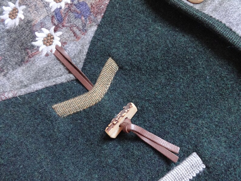 Puede incluir: Primer plano de una chaqueta de lana verde con cierre de bot&oacute;n de cuero marr&oacute;n. El bot&oacute;n est&aacute; hecho de asta de ciervo y tiene un patr&oacute;n decorativo. La chaqueta tiene un ribete de cuero marr&oacute;n y un tejido estampado en gris y blanco en el interior.